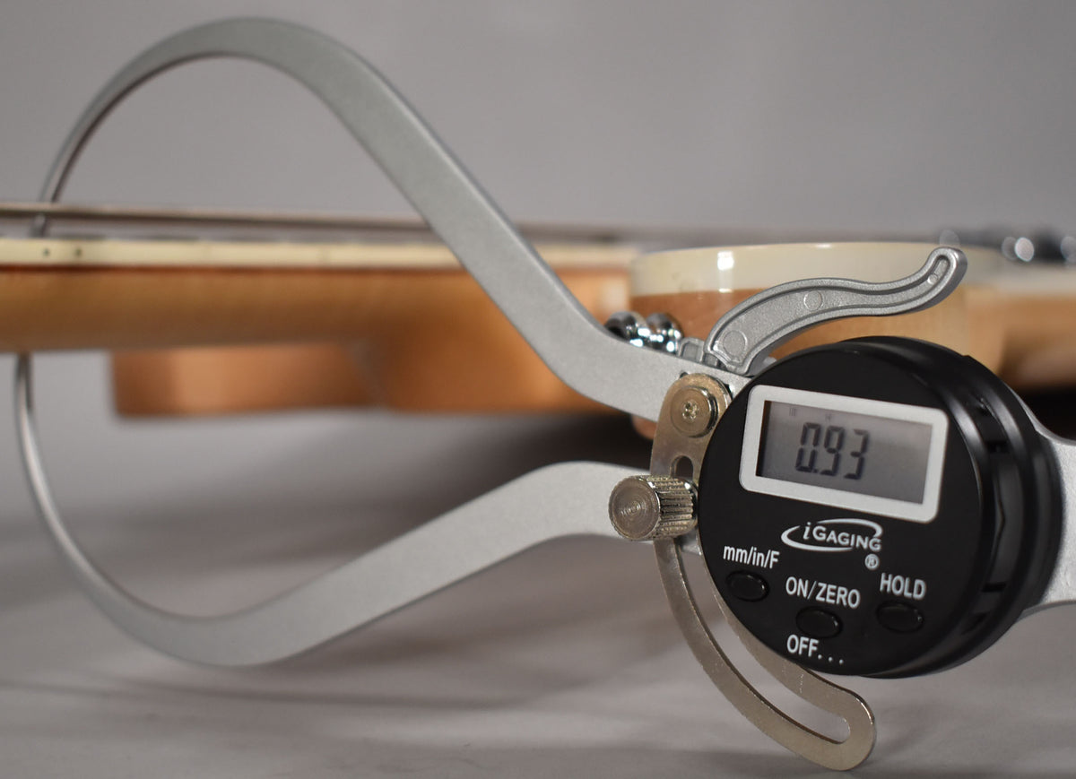 A digital caliper measures the string height of the bass, showing 0.93mm, with the light maple neck and body visible behind.