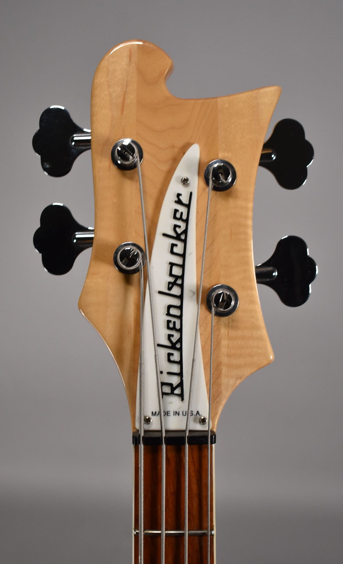 Close-up of the bass headstock, showing the natural maple finish, black clover-leaf tuners, and white truss rod cover.