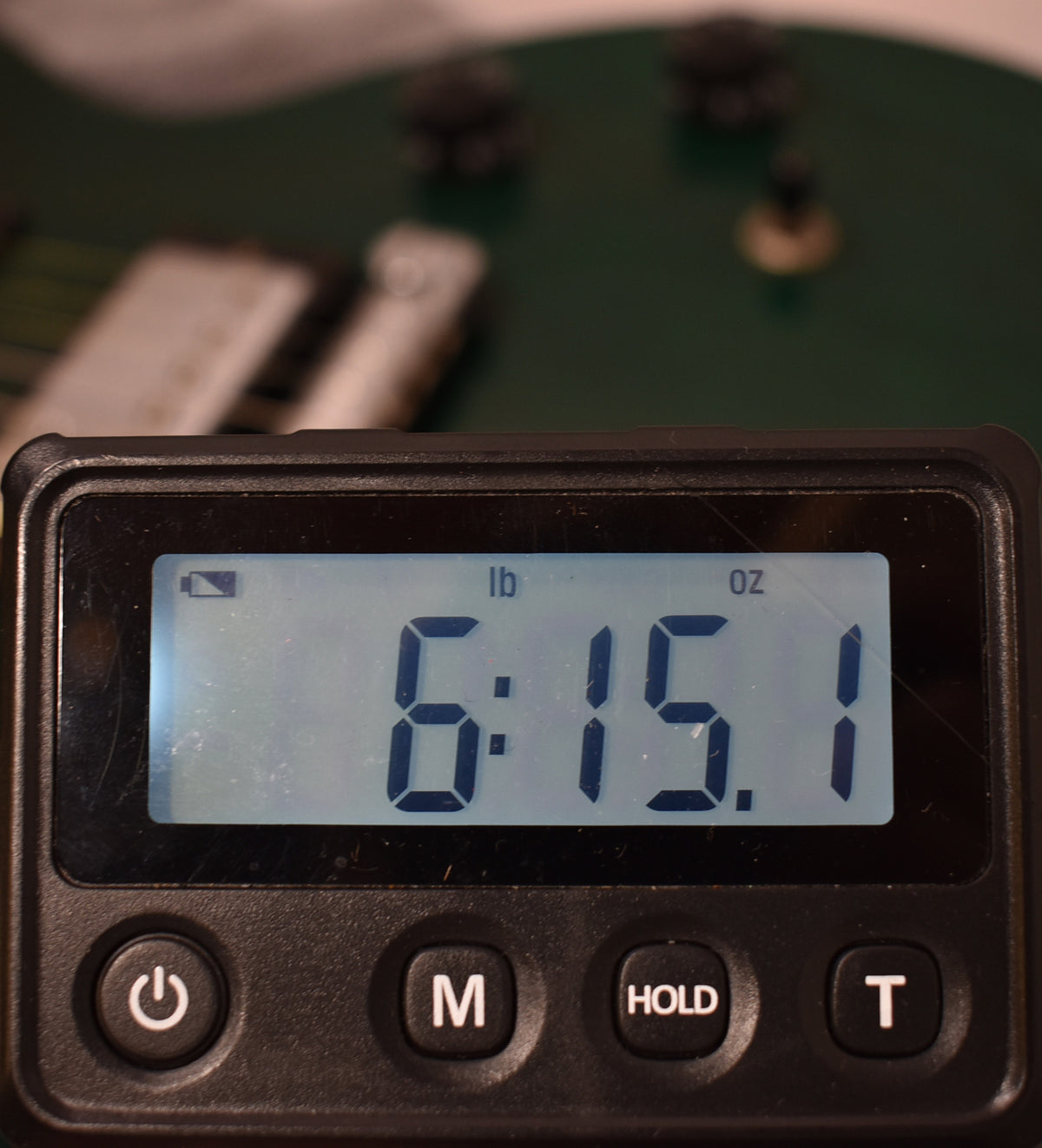 A digital scale displays "6:15.1" lbs and oz, with a blurred emerald green guitar body in the background.