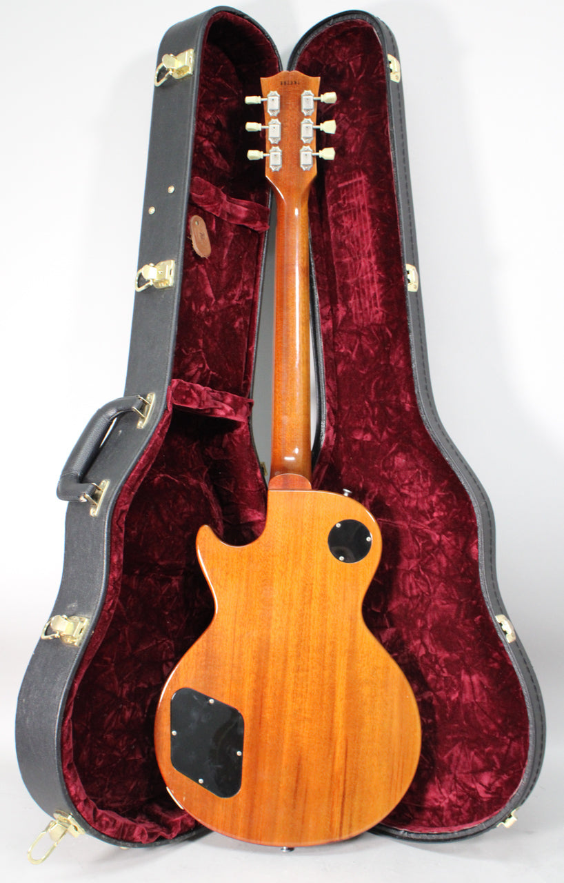 The guitar's back and neck are visible, a warm amber-brown with a glossy finish, resting in a black hard case with red velvet lining.