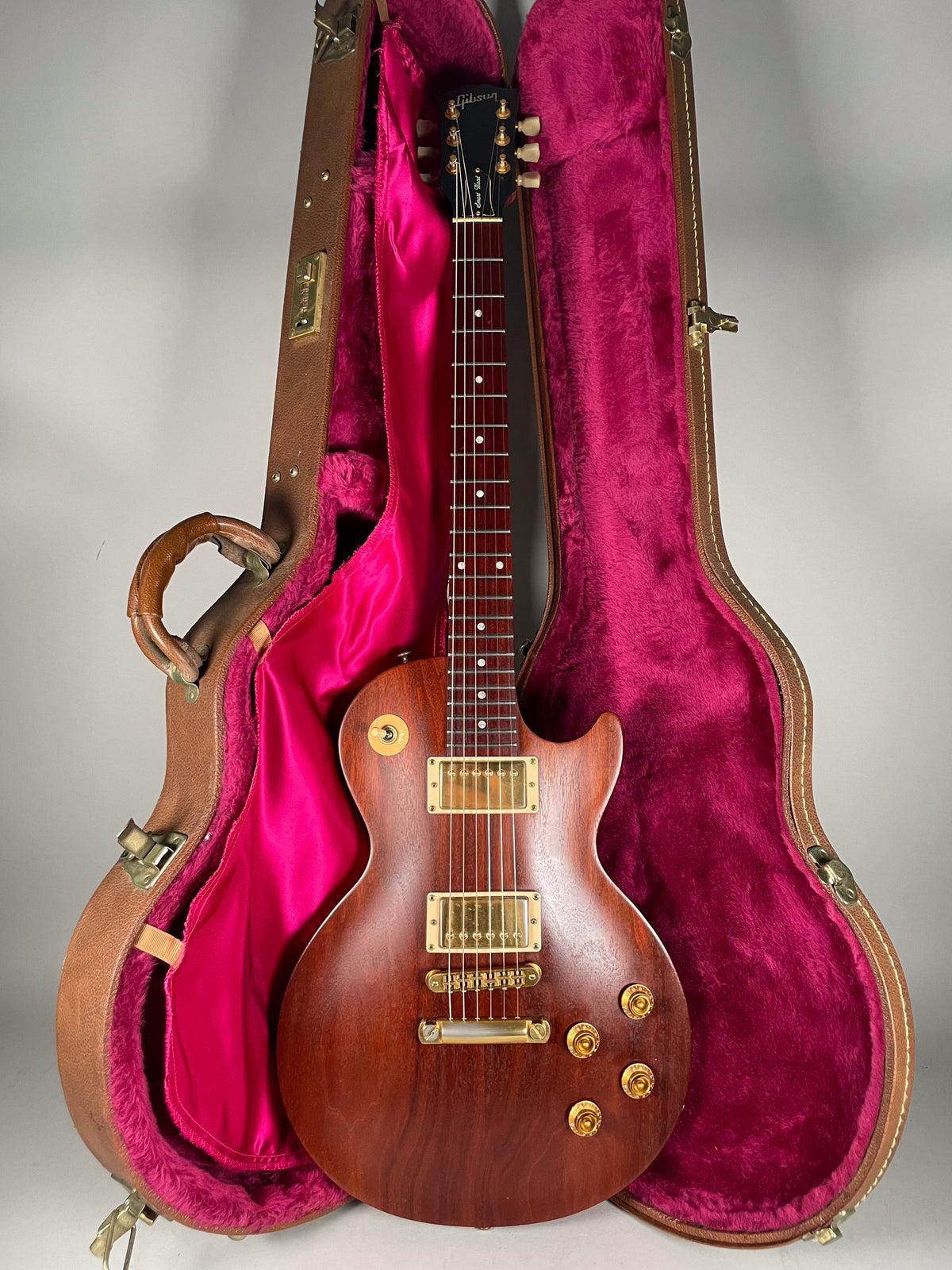 Full front view of a natural finish Les Paul with gold hardware, standing in a brown hard case with bright pink plush lining.