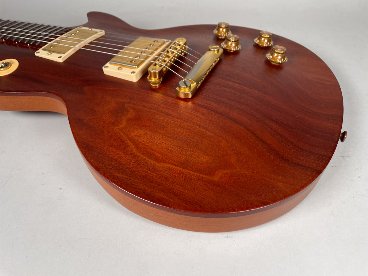Angled view of a reddish-brown guitar body with a natural wood grain finish, gold hardware, cream pickup rings, and four amber control knobs.