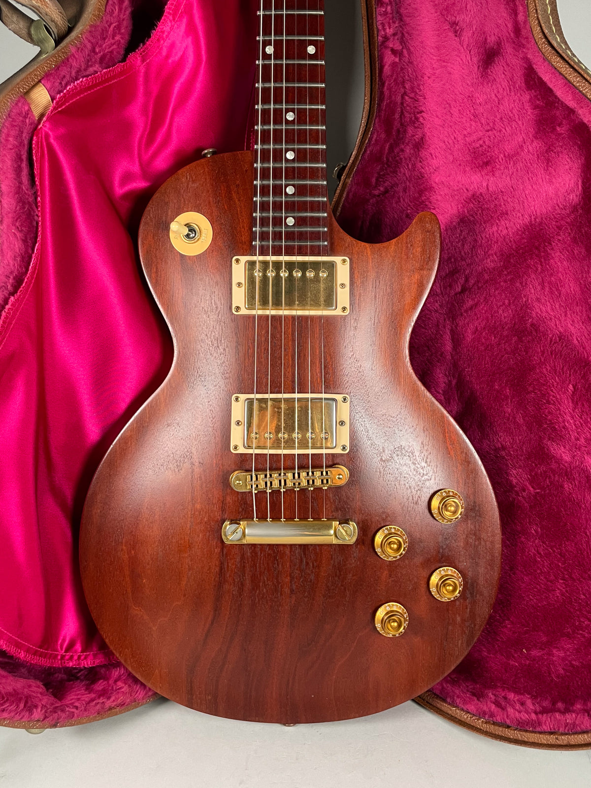 Full-frame view of a natural finish mahogany guitar with gold hardware, including two humbuckers and four control knobs, in a pink case.