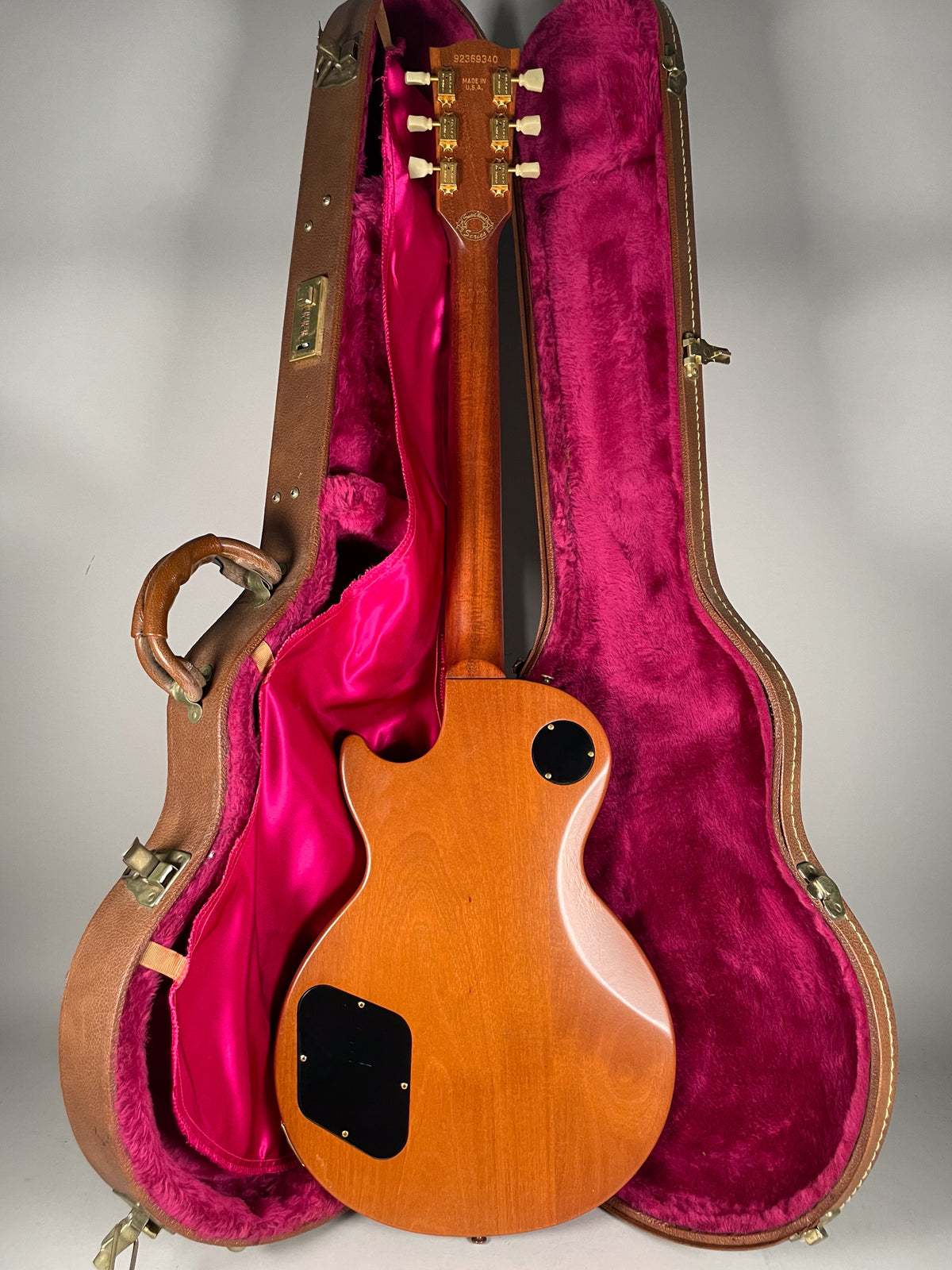 The guitar's natural wood back and neck are visible, with gold tuners and a black control cavity cover, resting in a pink-lined case.