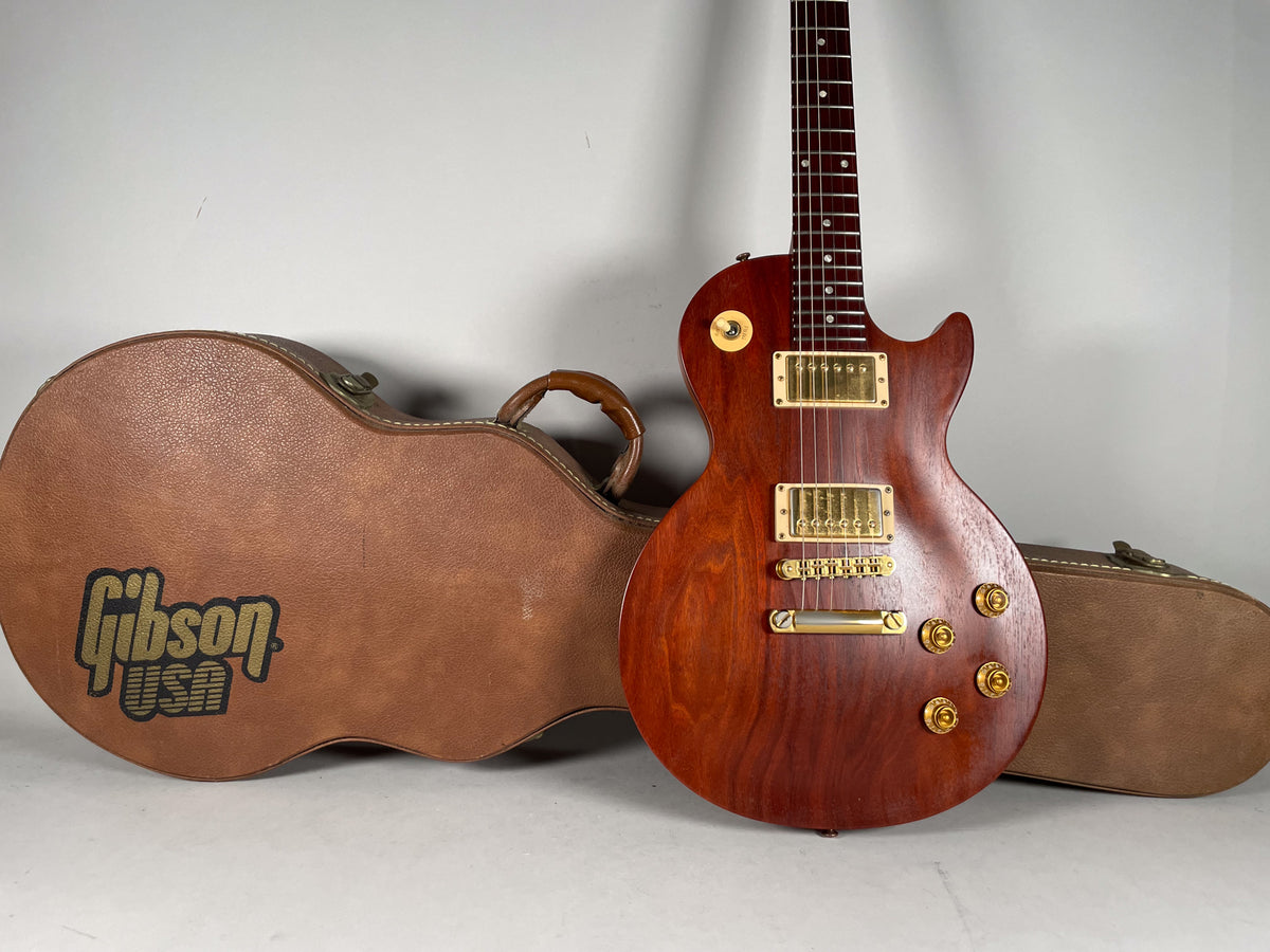 A natural finish Les Paul with gold hardware and four gold control knobs, standing next to its brown leather hard case.