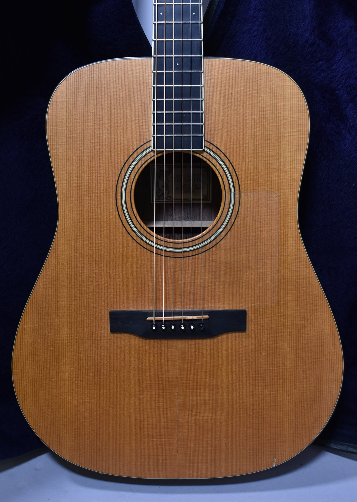 Full front view of a light brown acoustic guitar with a clear pickguard and black bridge, showing minor wear on the lower bout.