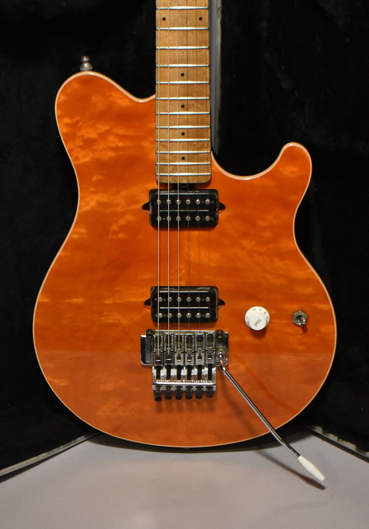 A close-up of an orange flame maple top guitar, showing the neck, two black humbuckers, a chrome Floyd Rose tremolo, and white volume knob.