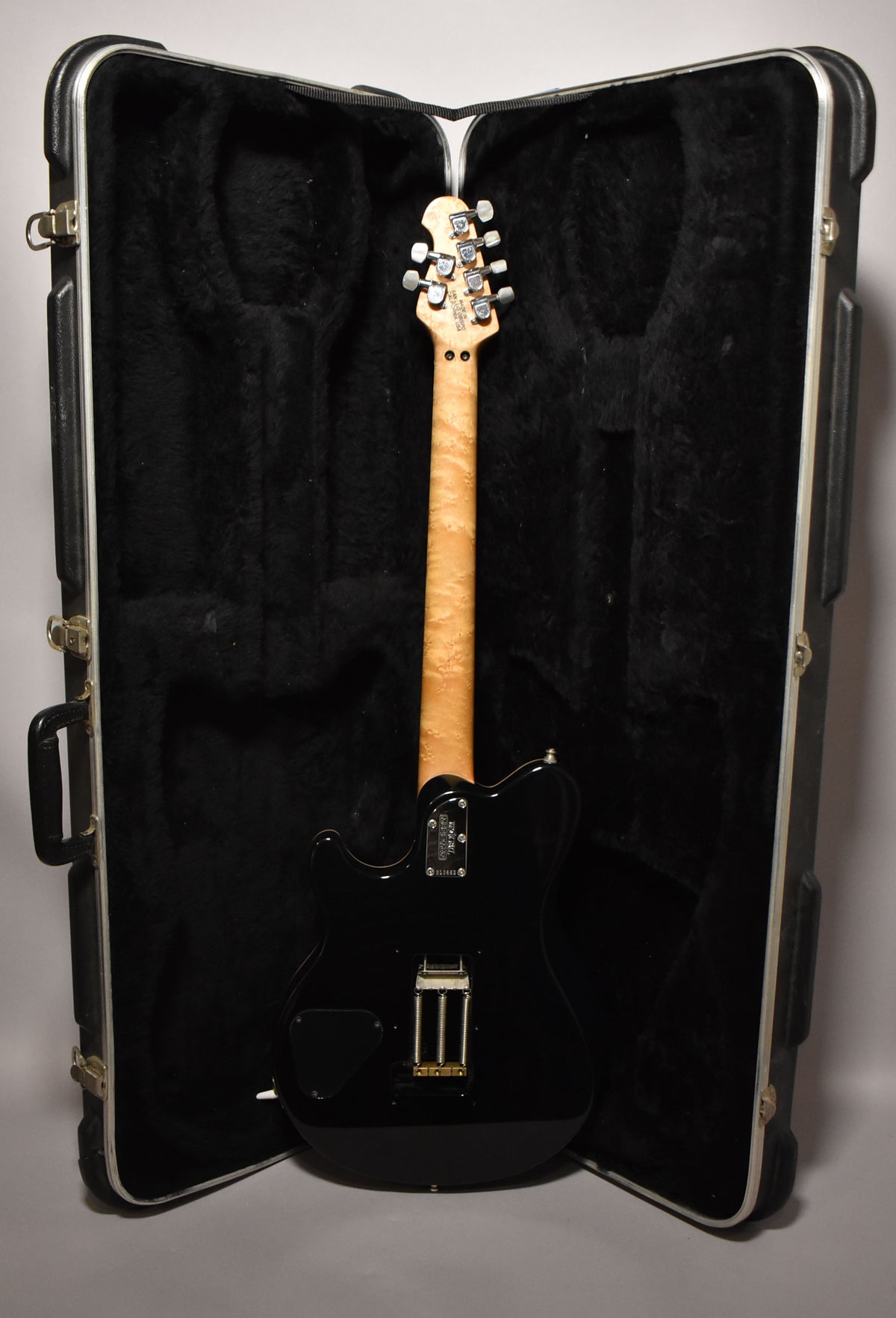 The black solidbody guitar is shown from the back, resting in its open hard case, revealing a flame maple neck and headstock.