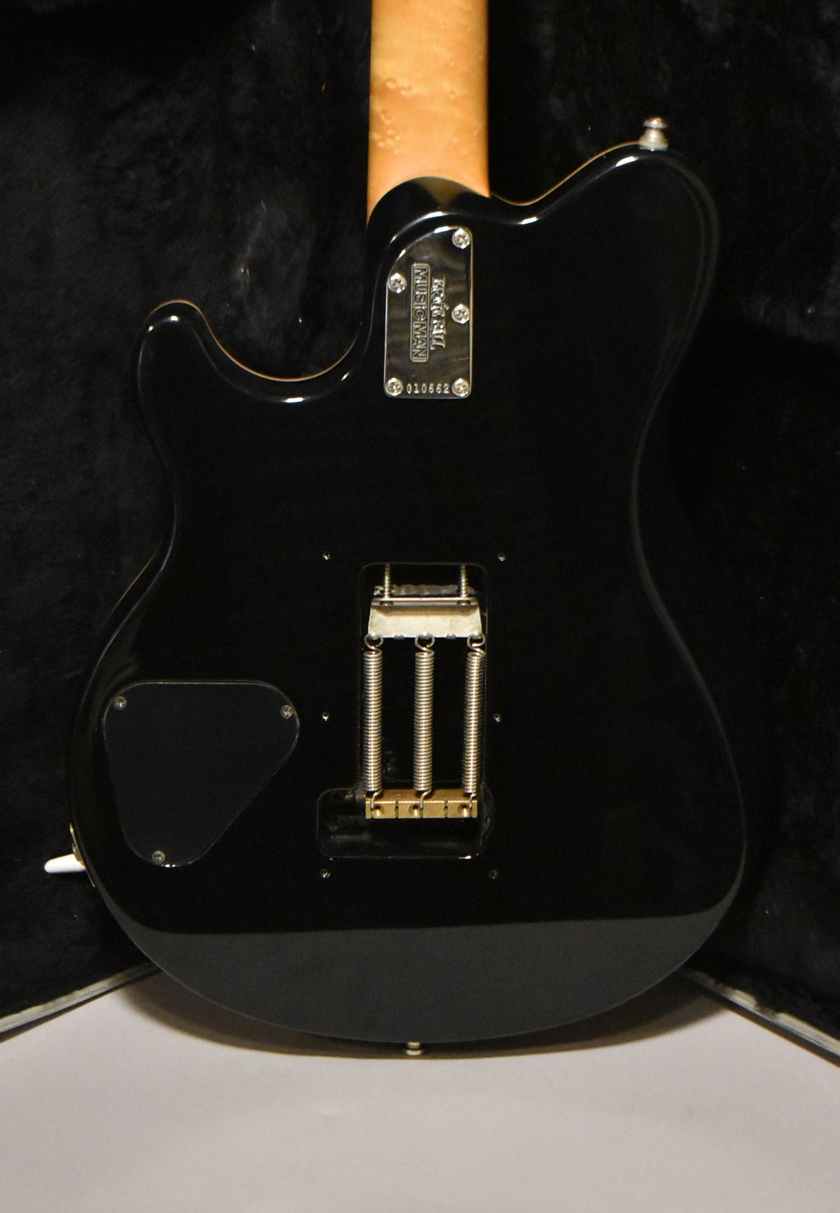 The back of a glossy black solidbody electric guitar, showing a natural maple neck, gold tremolo block, and black control cavity cover.