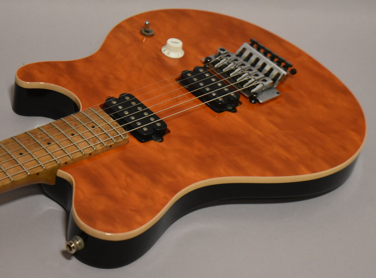 The guitar's body, with a flame maple top in amber, black sides, and cream binding, is angled to show the tremolo and pickups.