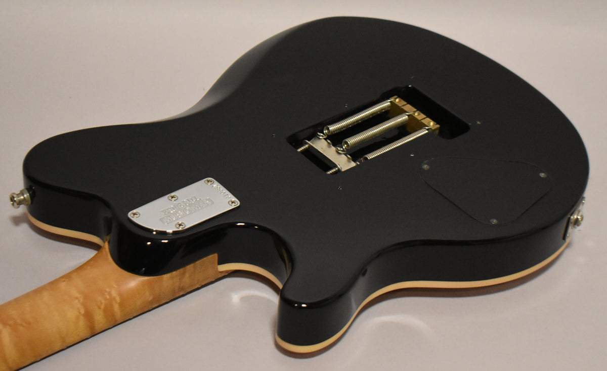 The guitar's glossy black back shows a light maple neck, a chrome neck plate, and an open tremolo spring cavity.