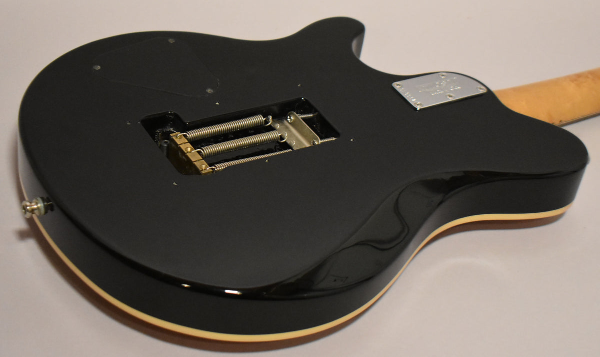 The back of a black solidbody guitar, showing the tremolo spring cavity with gold hardware, a chrome neck plate, and cream binding.