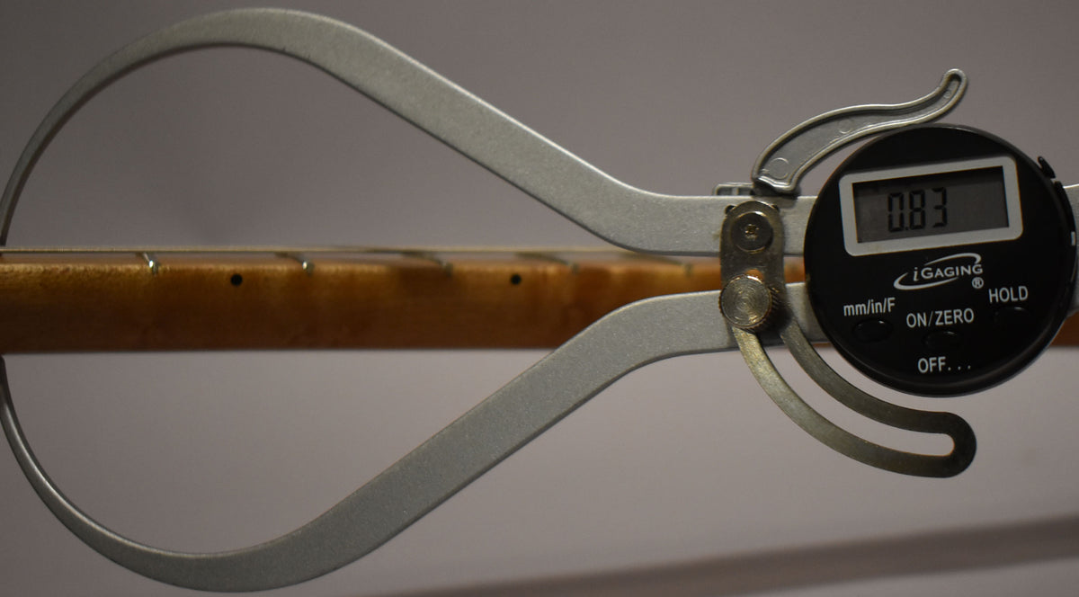 A digital caliper measures the guitar's fret height at 0.83mm, showing the light brown maple neck and dark fret markers.