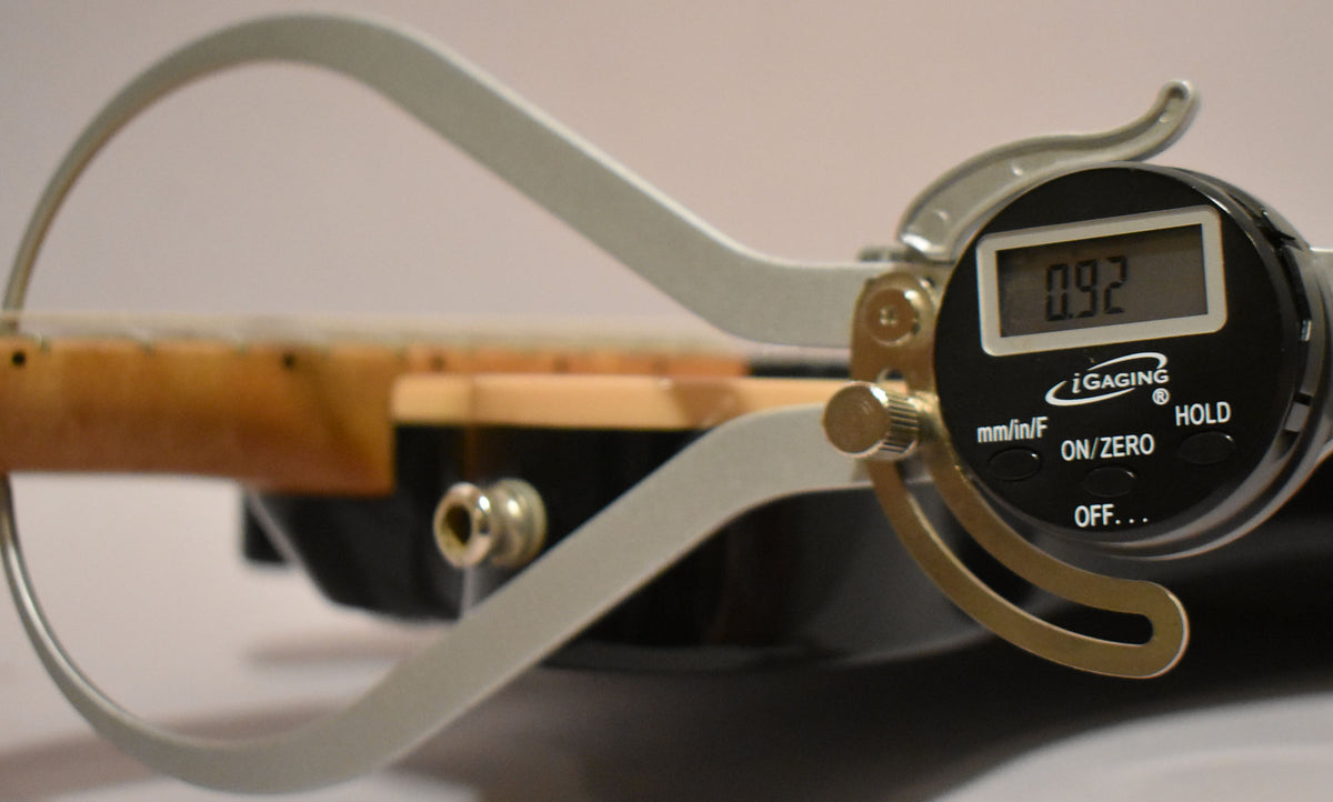 A digital caliper measures the neck relief of the guitar, showing 0.92mm, with the maple neck and black body visible.