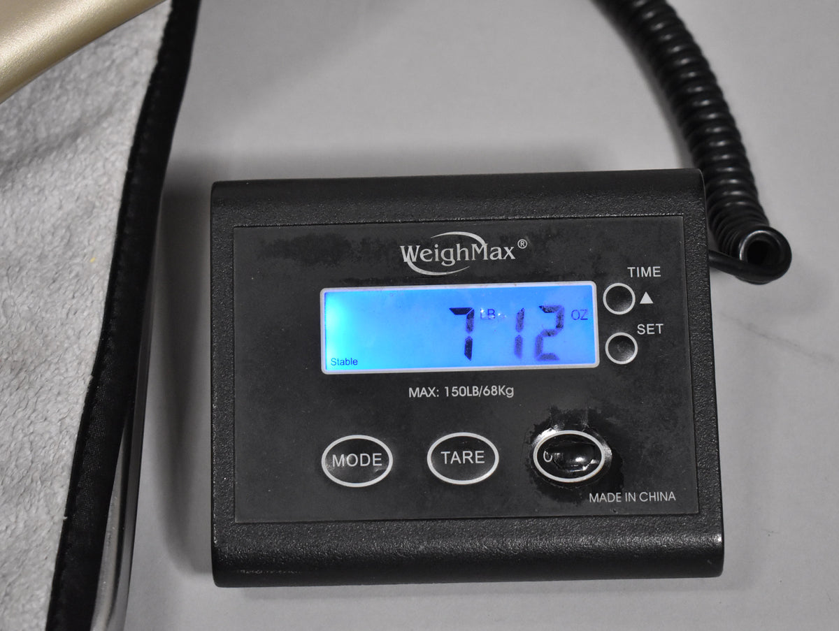 A close-up of a black digital scale displaying "7 LB. 12 OZ." on a blue screen, with part of a guitar case visible on the left.