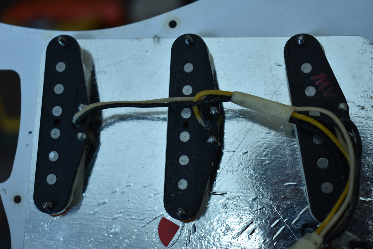 Close-up of three black single-coil pickups with white pole pieces, showing wiring and a scratched silver shielding plate.