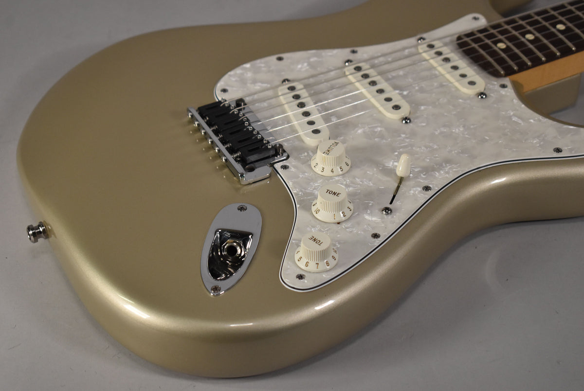 Upper body of a metallic gold Stratocaster, angled right, showing a pearloid pickguard, three cream single-coil pickups, and three cream control knobs.