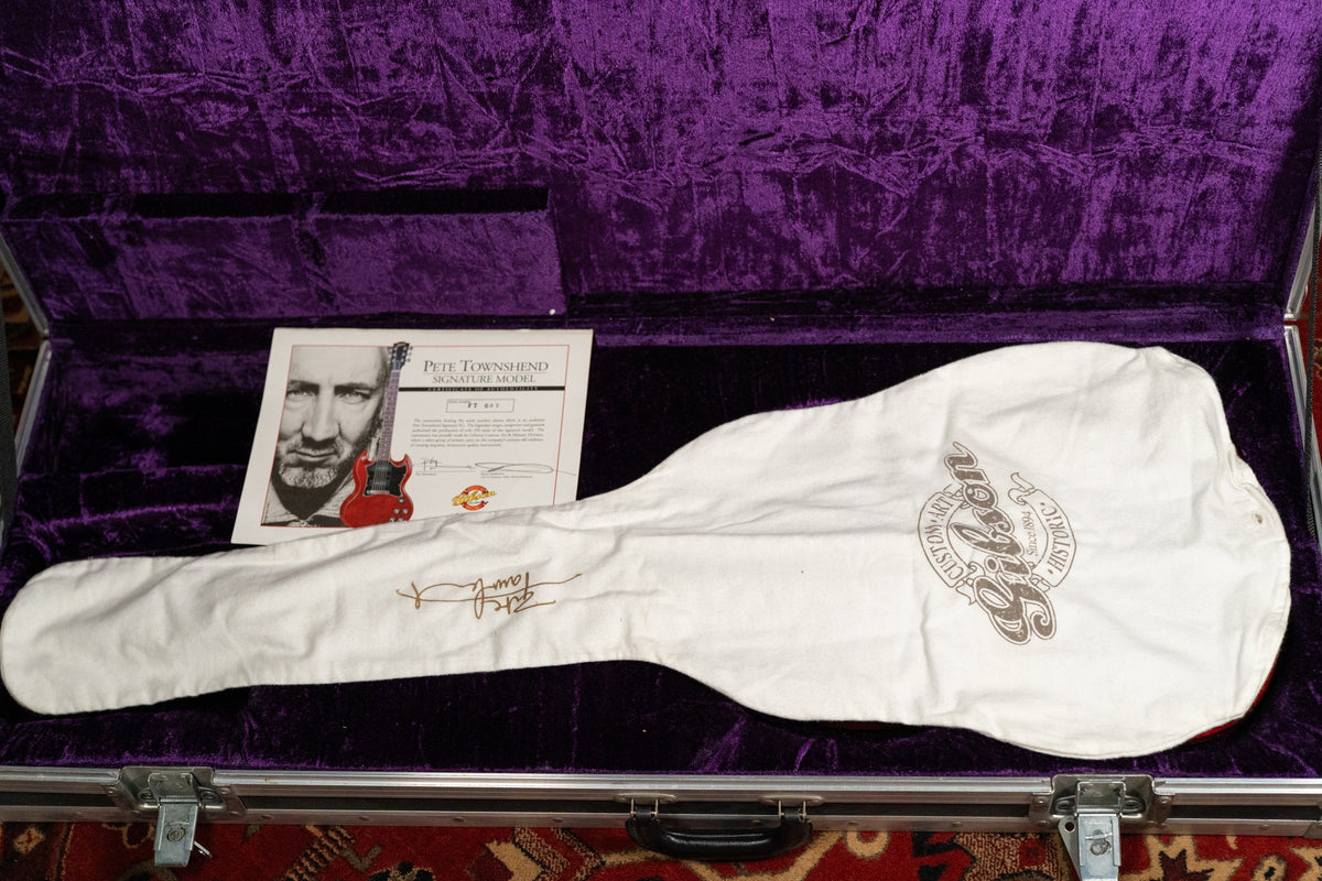 The guitar, covered by a white cloth bag, rests in a purple velvet-lined hard case, with a certificate of authenticity visible.