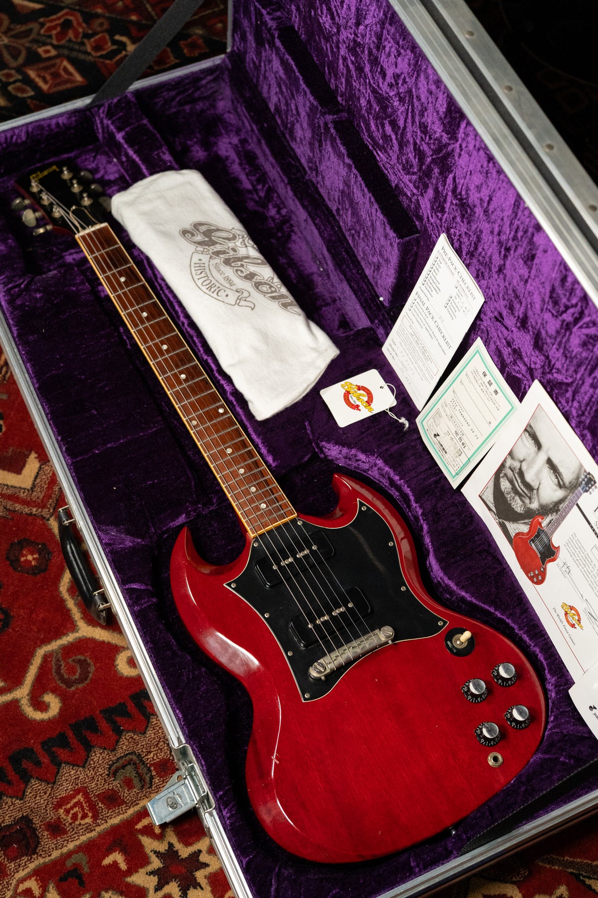 A cherry red SG Special with a black pickguard and P90 pickups rests in a purple-lined case with papers and a white cloth.