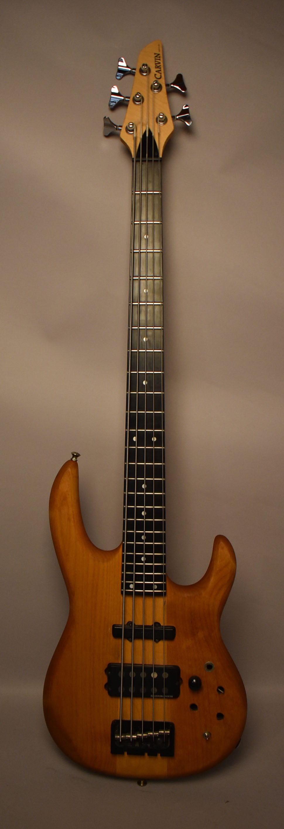 Full-length view of a natural wood 5-string bass with black pickups and bridge, chrome tuners, and several empty control holes.