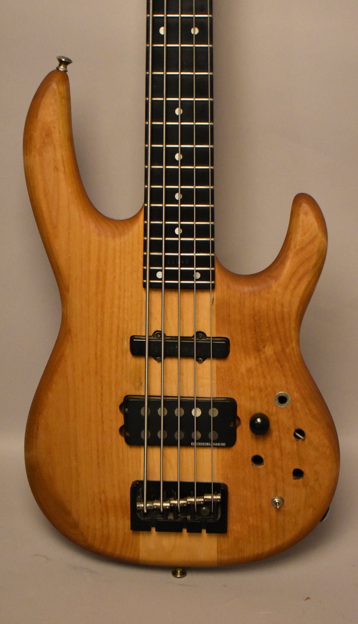 Full-frame view of a natural wood bass with a dark fretboard, two black pickups, a black bridge, and missing control knobs.