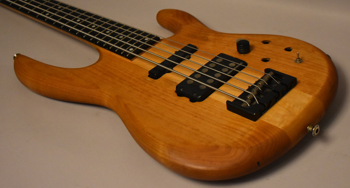 A natural wood 5-string bass guitar, with black pickups and bridge, angled from the neck to the lower bout.