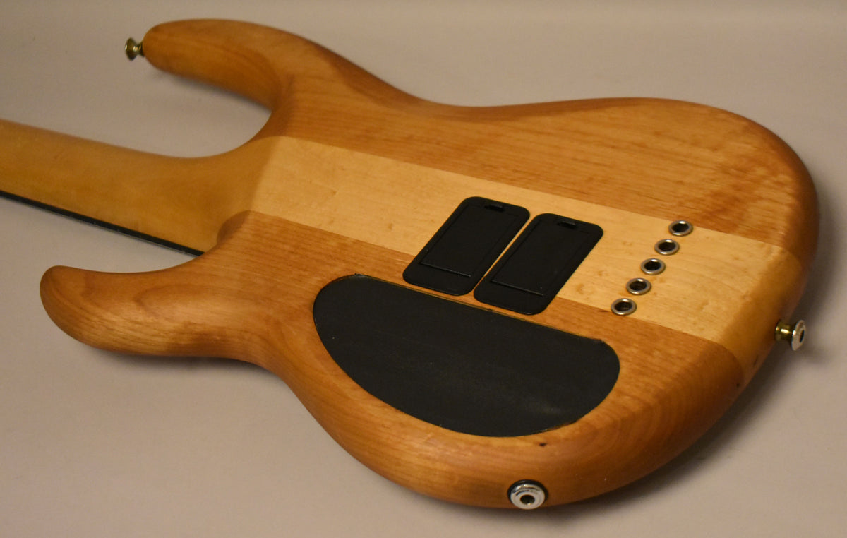 The back of a stripped natural wood bass body, showing the neck joint, two battery covers, and five string ferrules.