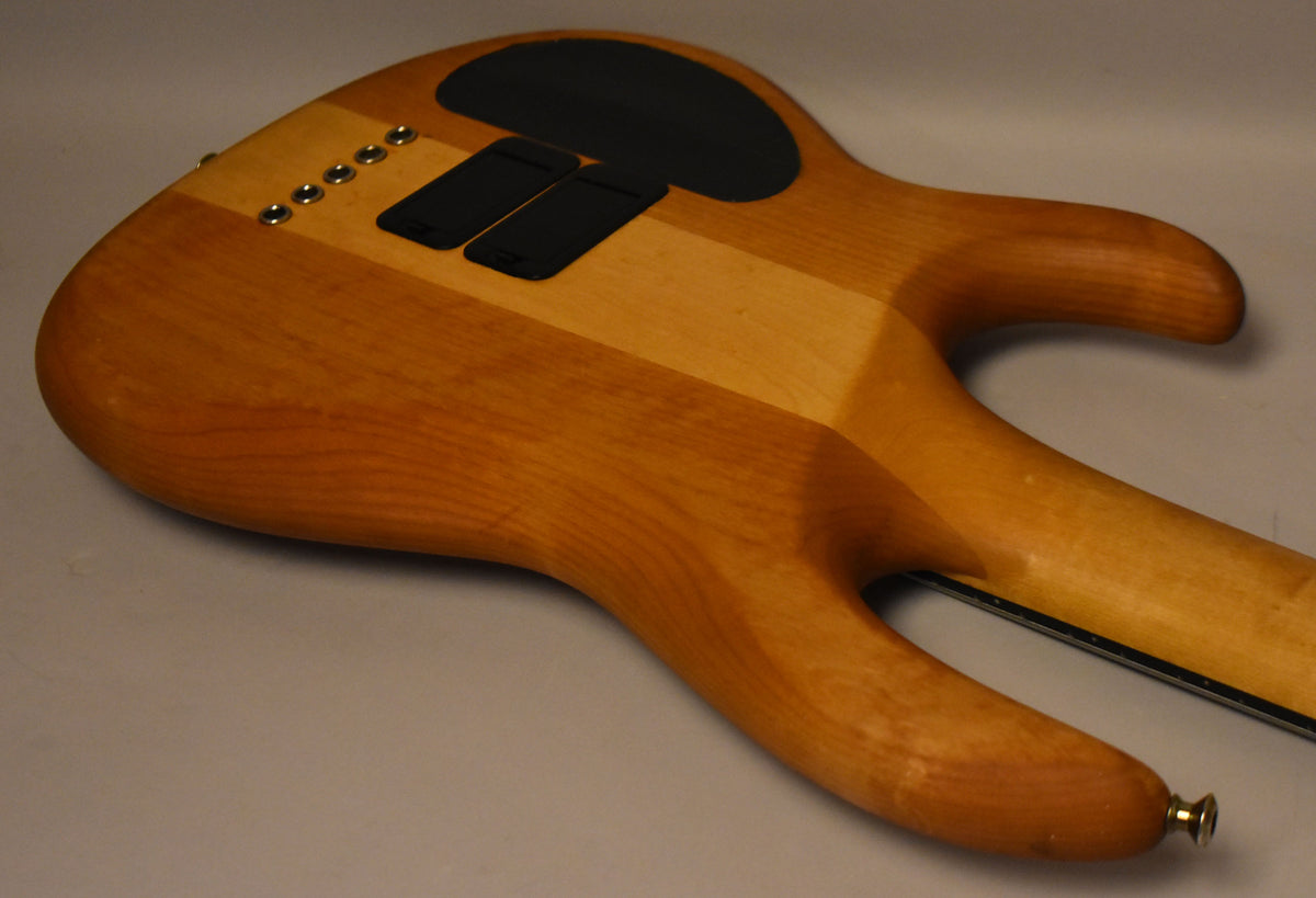 The back of a light brown bass body with darker wood stripes, showing five string ferrules and three black control cavity covers.
