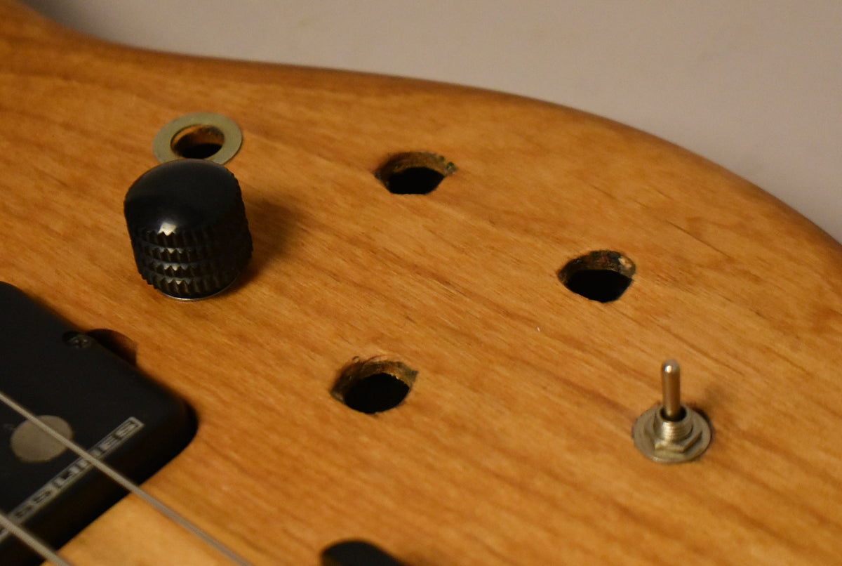 Close-up of the bass's natural wood body, showing a black knob, a chrome toggle switch, and three empty control holes.