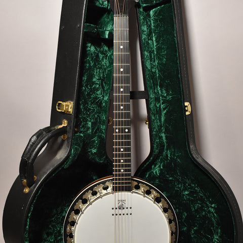 A black 6-string banjo with a white head and gold hardware, resting in its open black hard case with a dark green crushed velvet interior.