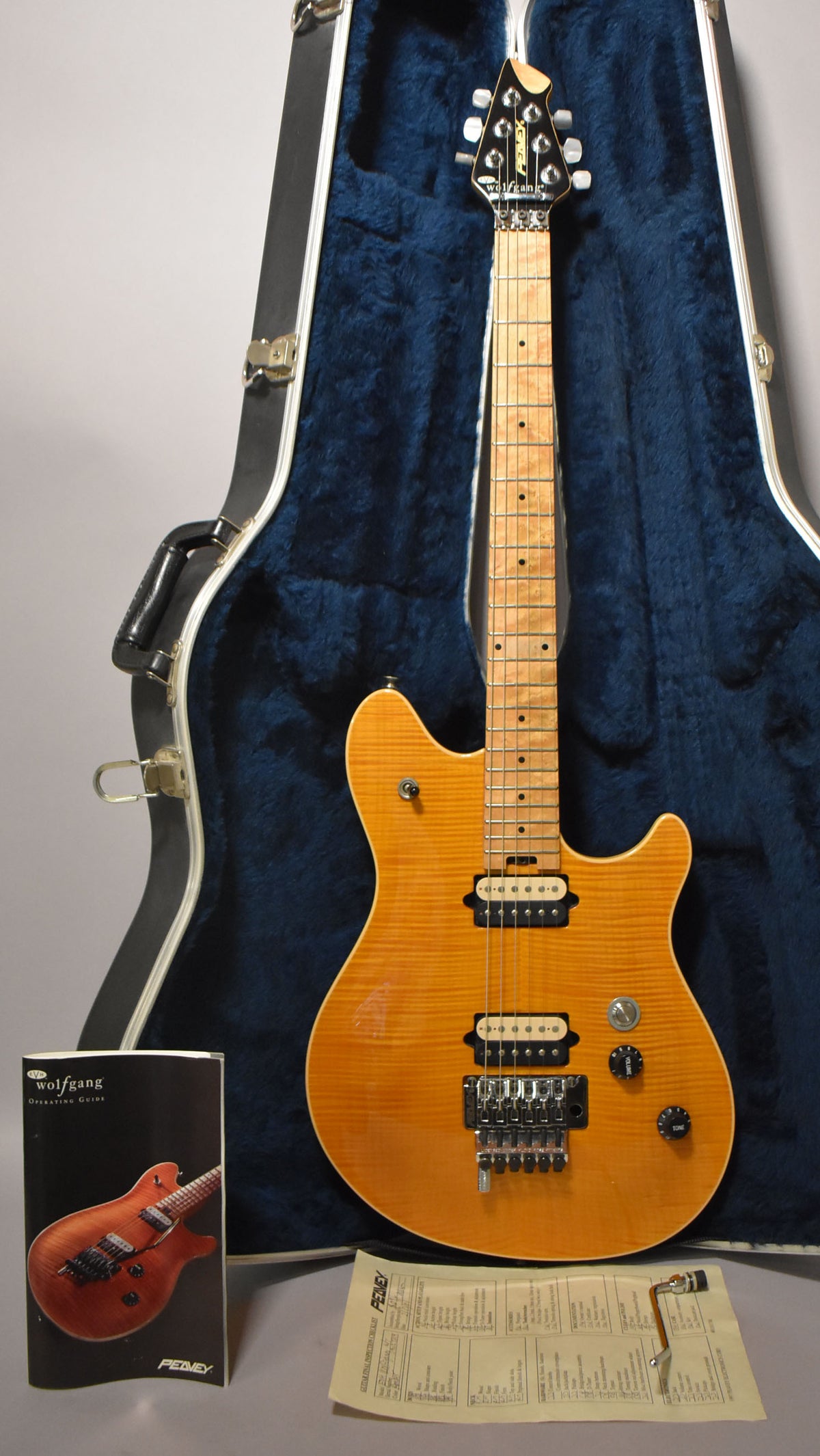 Full-length view of an amber flame-top guitar with a maple neck, black headstock, and chrome tremolo system, resting in a blue-lined case.