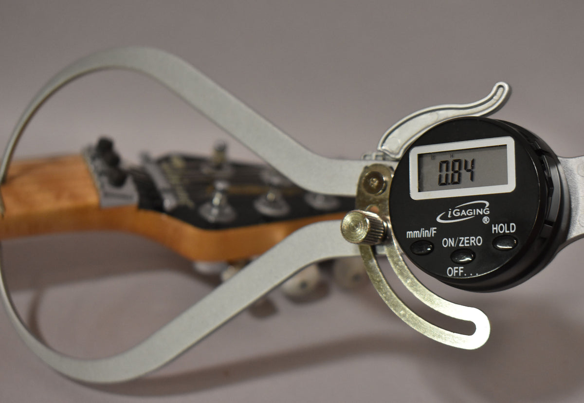 A digital caliper measures the guitar's amber-finished neck, showing a reading of 0.84, with tuning pegs visible in the background.