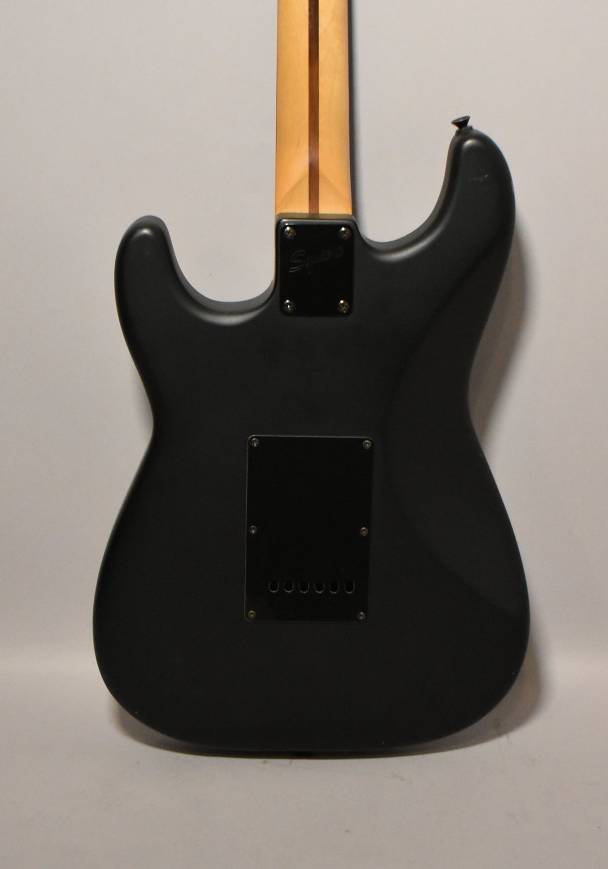 The back of a satin black solid body electric guitar, showing the neck plate, tremolo cavity cover, and string-through-body ferrules.