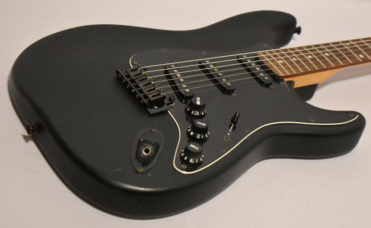 The black satin Stratocaster body is angled, showing the black pickguard, three single-coil pickups, and three control knobs.