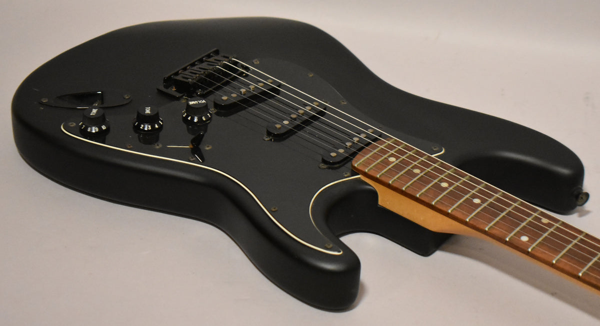 Upper body of a black satin Stratocaster, angled from the lower bout, showing the black pickguard, three pickups, and rosewood fretboard.