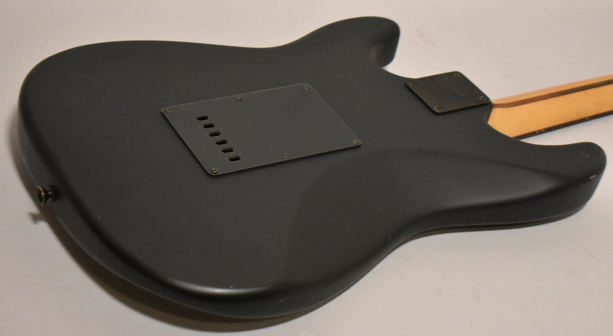 The back of a satin black solid body guitar, angled from the lower left, showing the tremolo cavity cover and neck plate.