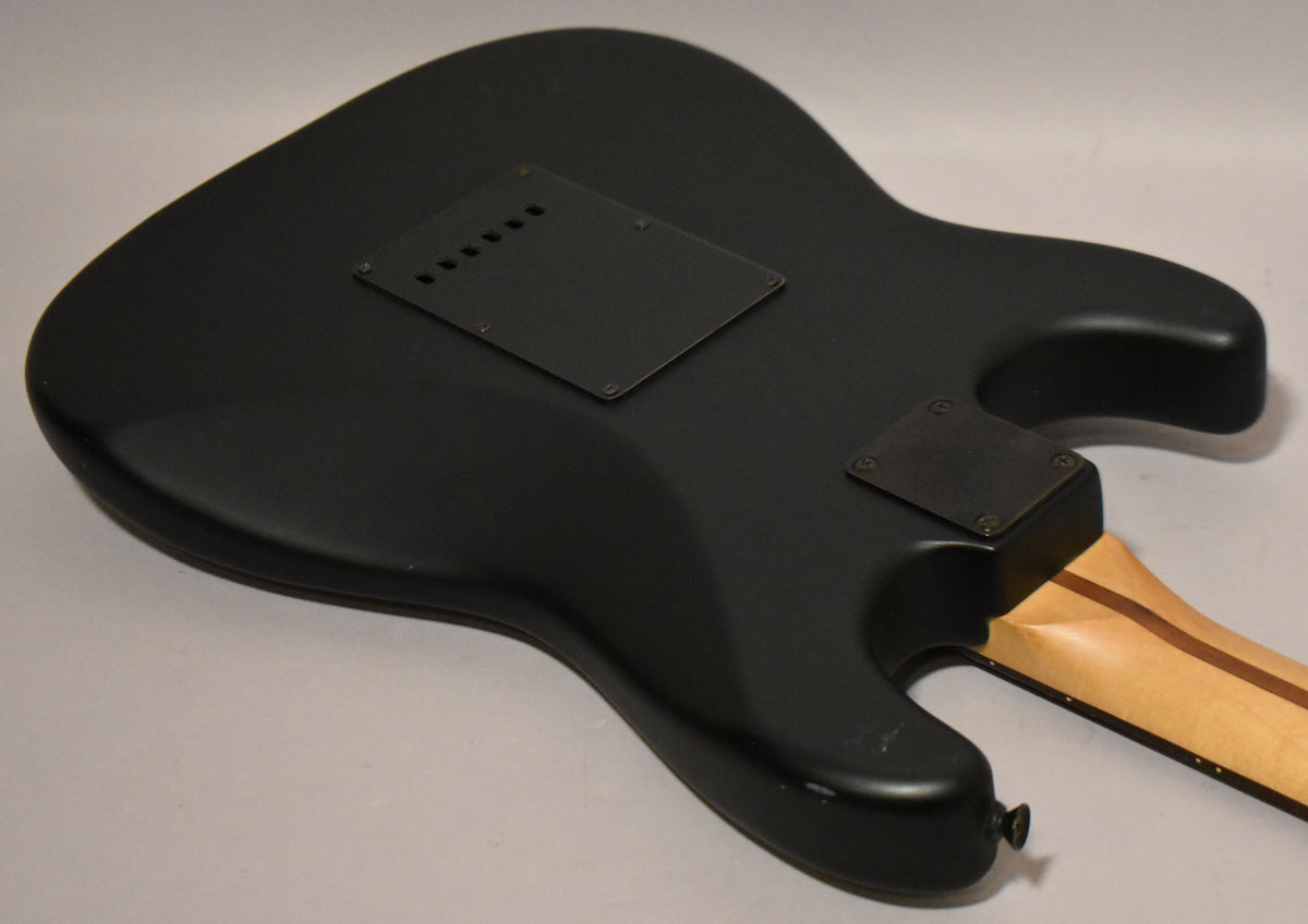 The satin black guitar body is seen from the back, angled right, revealing two control plates and a light wood neck.