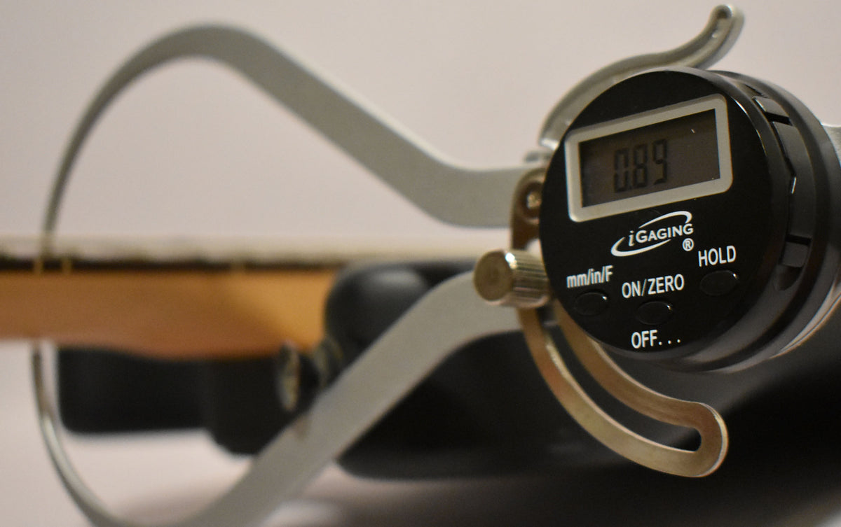 A digital caliper measures the neck relief of the guitar, displaying "0.89" on its screen, with the neck blurred in the background.