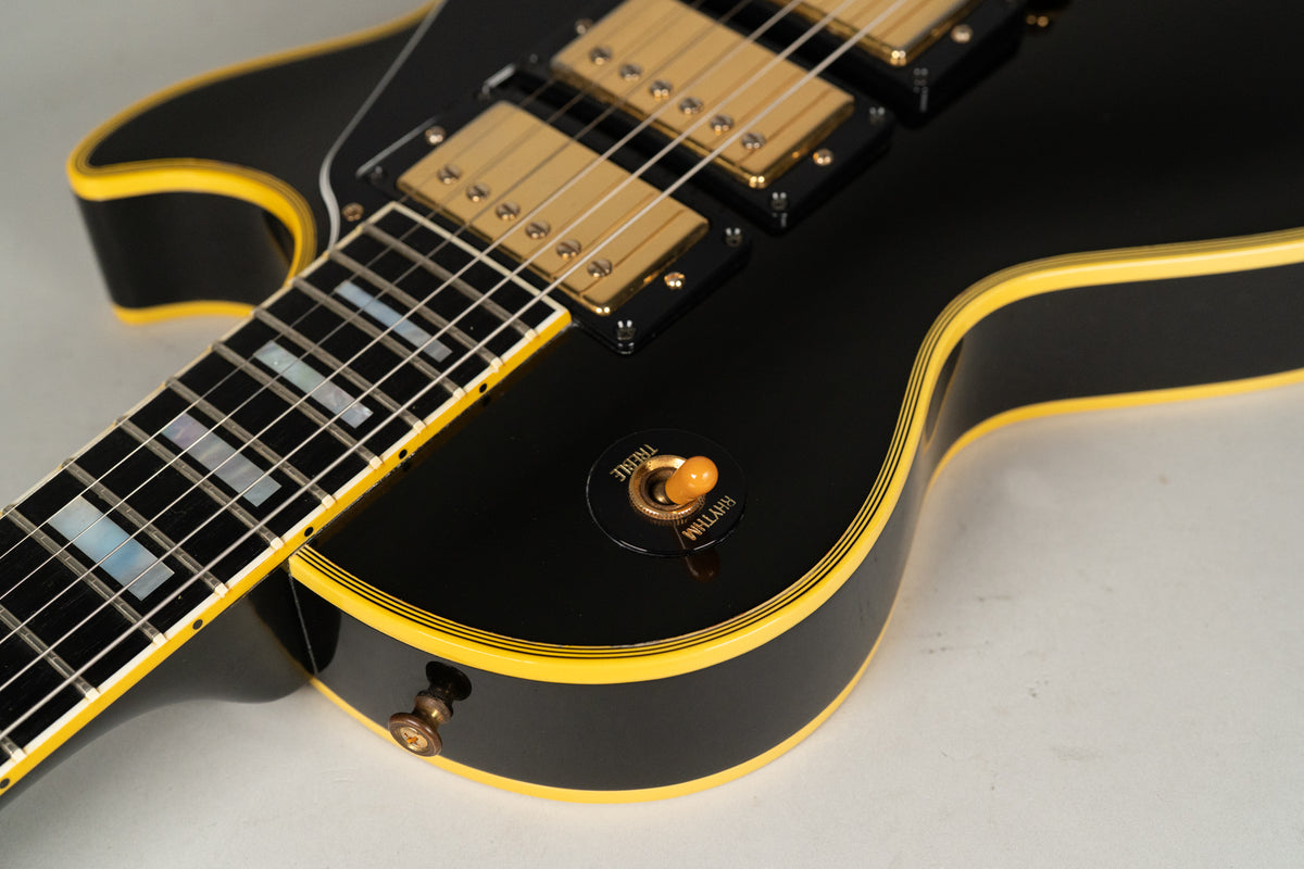 Close-up of the black guitar body with yellow binding, showing gold pickups, an amber switch tip, and rectangular fretboard inlays.