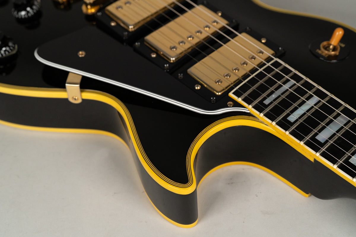 Close-up of a black electric guitar body with multi-ply yellow binding, gold hardware, and a dark fretboard with block inlays.