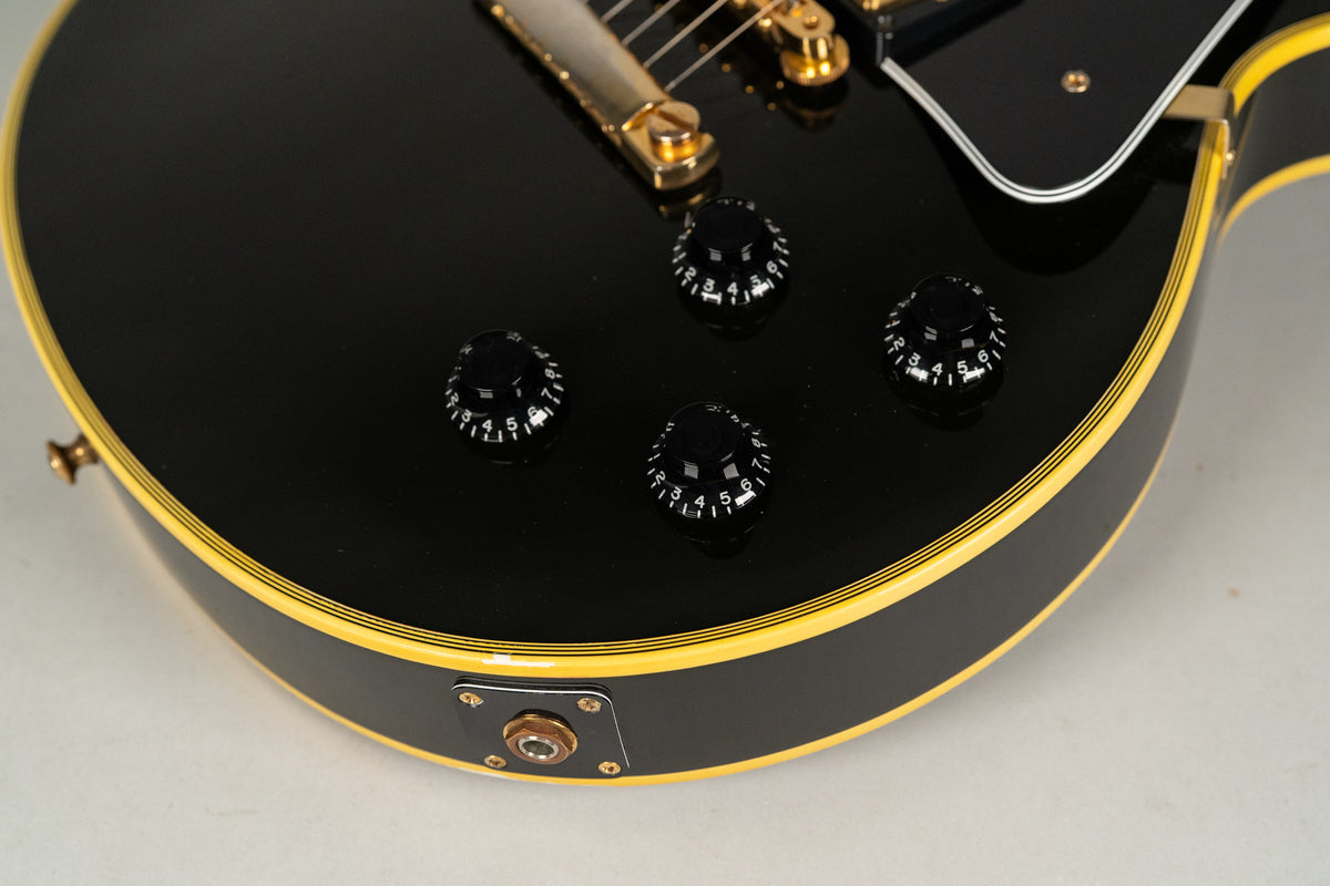 Close-up of the glossy black guitar body, showing four black numbered control knobs, a gold output jack, and multi-ply binding.