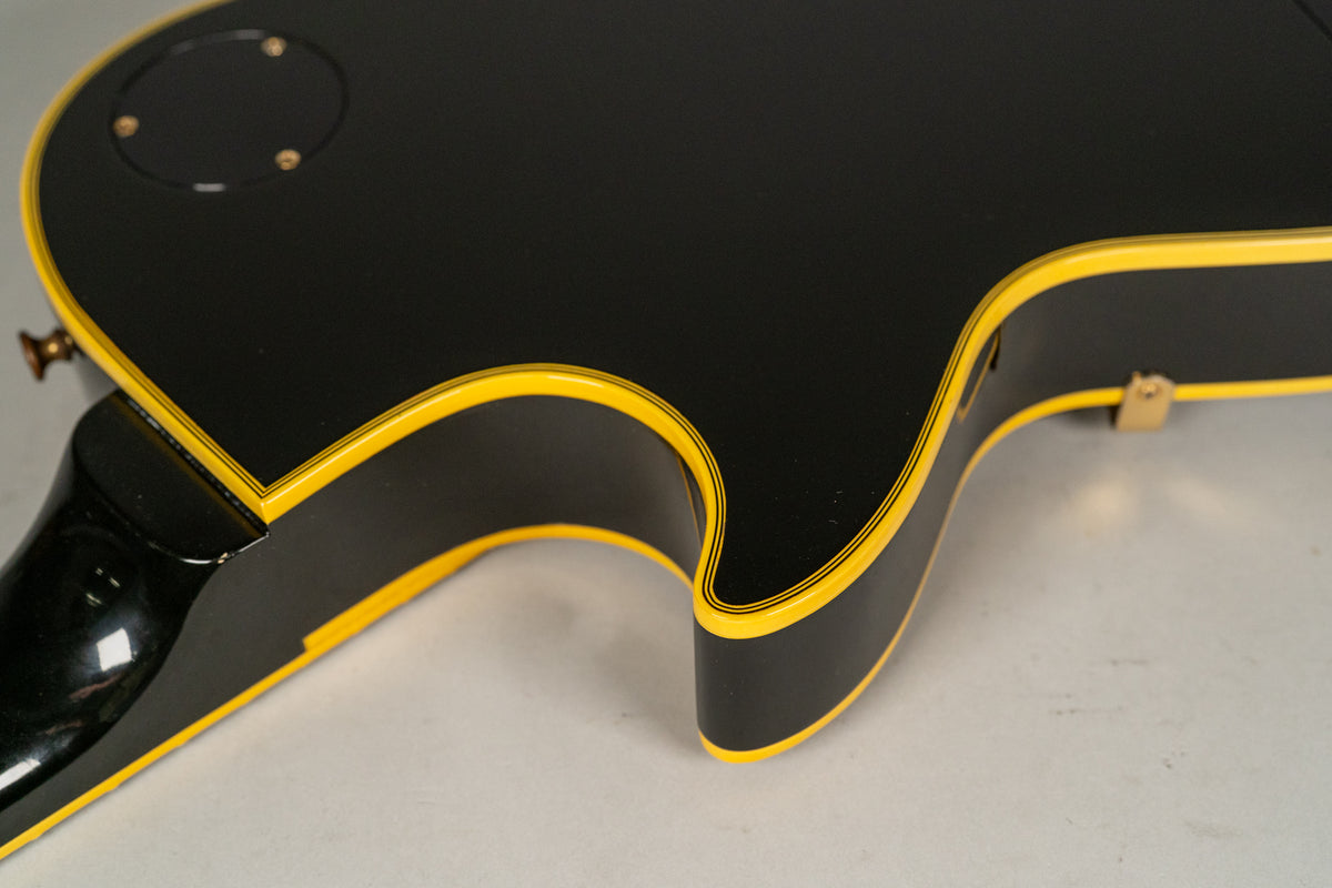The guitar's black body with yellow binding and pinstripes is shown from the back, angled to reveal the neck joint and control cavity cover.