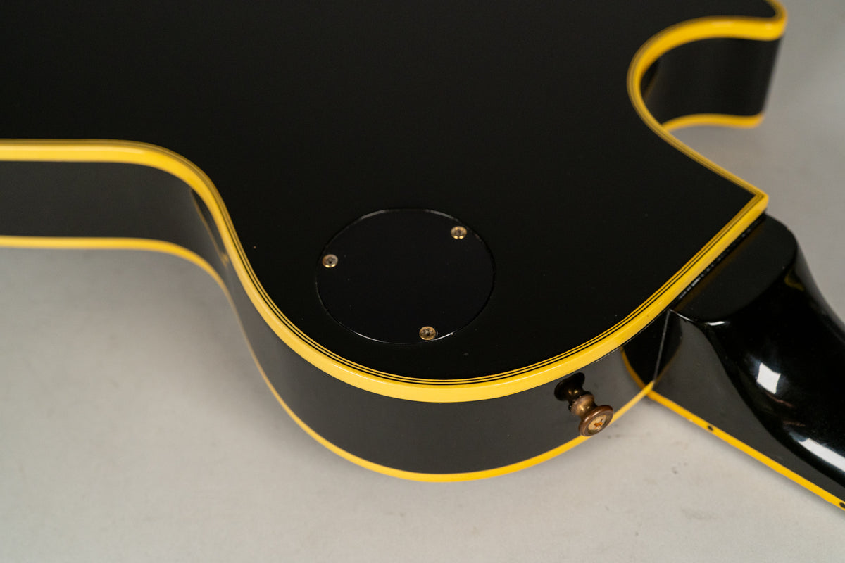The lower bout of a black guitar with yellow binding, showing the back control plate and a worn strap button.