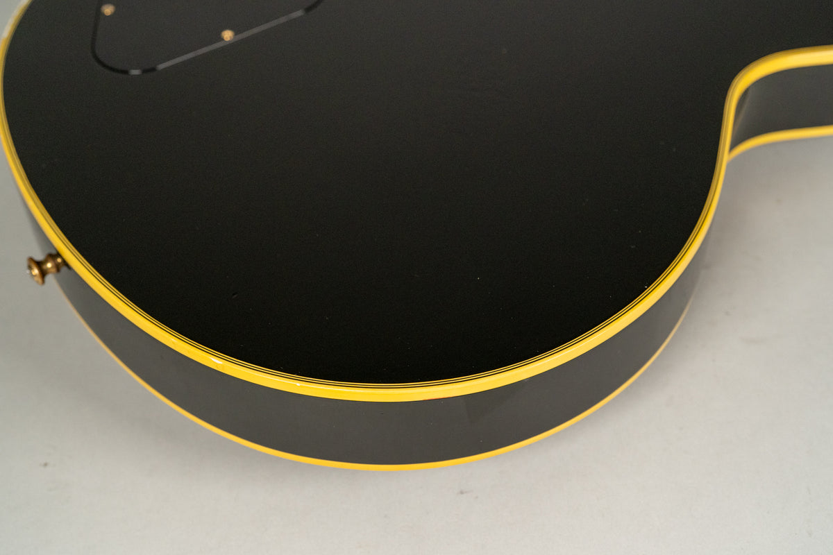Detail of a black guitar body with yellow binding and a gold strap button.