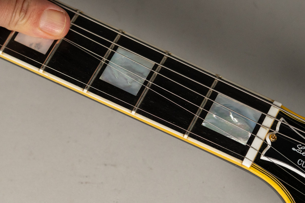 Close-up of the black fretboard with rectangular mother-of-pearl inlays, showing a thumb pressing the first fret.
