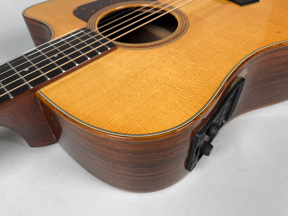 Upper bout of a natural finish acoustic guitar, showing the cutaway, dark wood sides, and black preamp controls.