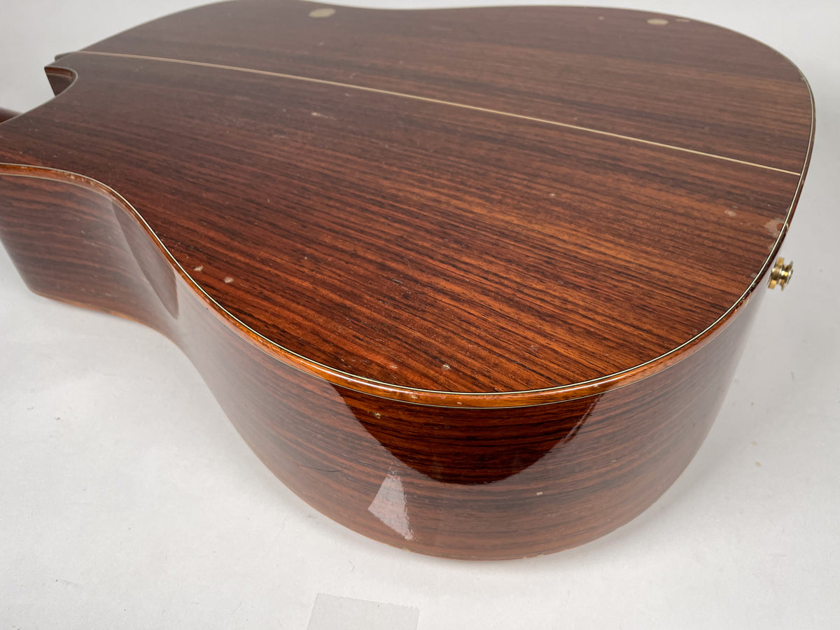 The guitar's dark wood back and side, with a cutaway, show light scratches and dings, and a gold endpin jack.