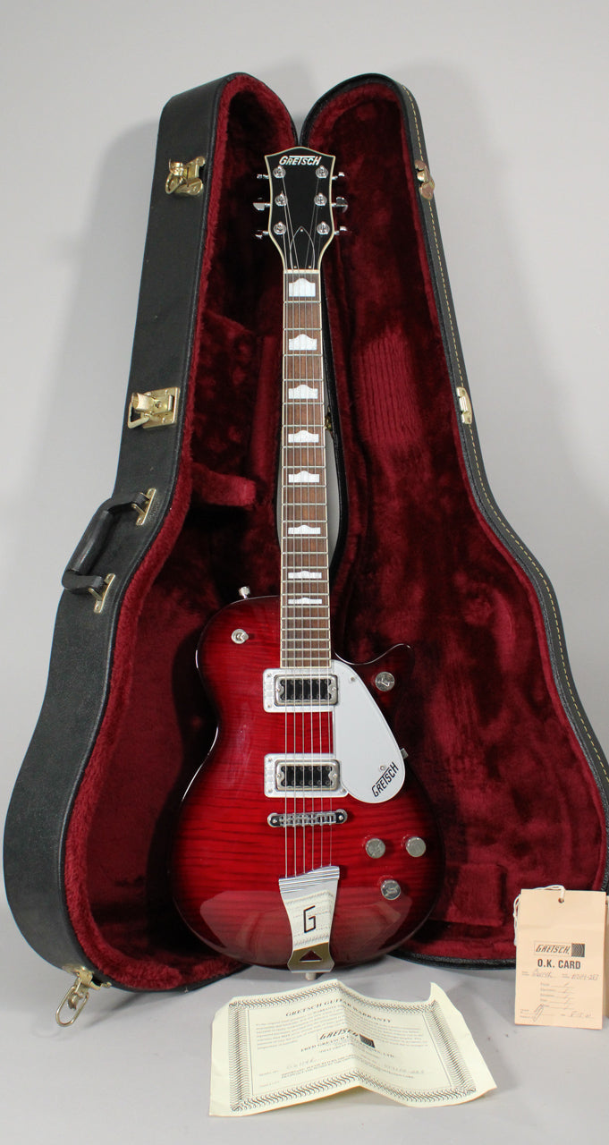 A transparent red Gretsch New Jet with a flamed maple top, white pickguard, and chrome hardware rests in its open, red-lined hard case.