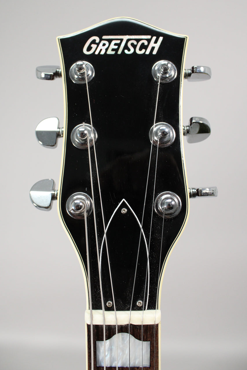 Close-up of the guitar's glossy black headstock with chrome tuners, white binding, and mother-of-pearl inlay, showing light dust.