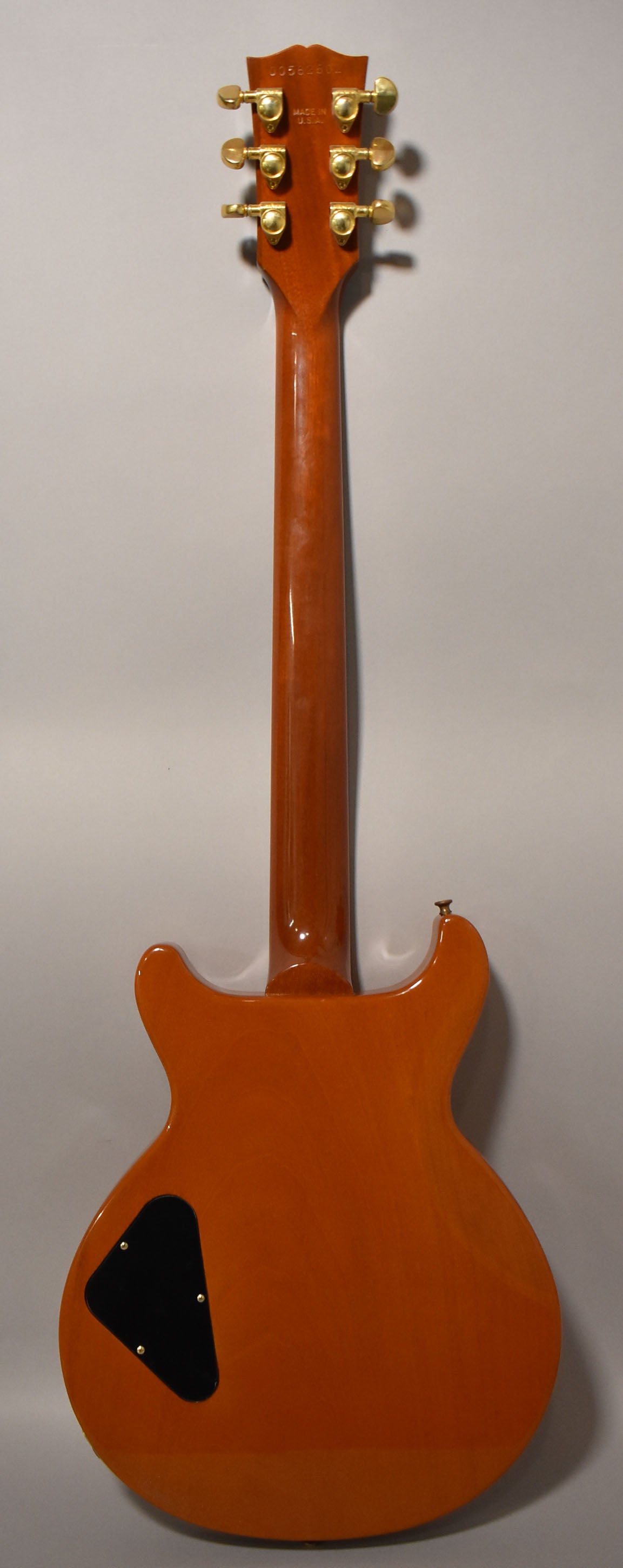 The back of a glossy amber guitar with a black control cavity cover and gold tuners on the headstock.